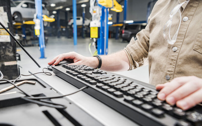 Caucasian Automotive Technician Checking Vehicle Database Documentation. Hands on Computer Keyboard Inside Auto Service Station.
