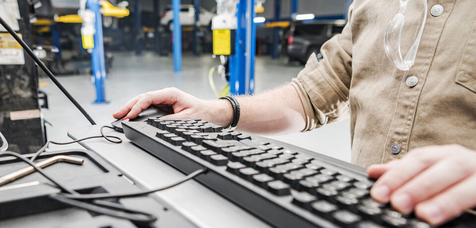 Caucasian Automotive Technician Checking Vehicle Database Documentation. Hands on Computer Keyboard Inside Auto Service Station.