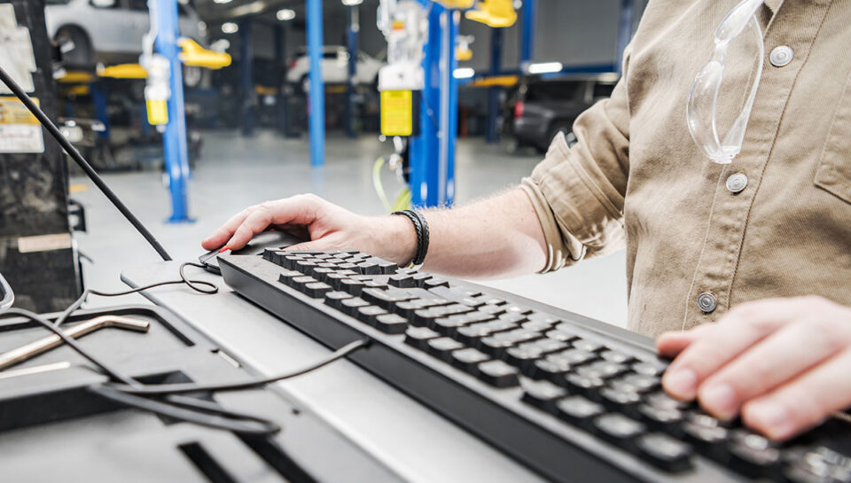 Caucasian Automotive Technician Checking Vehicle Database Documentation. Hands on Computer Keyboard Inside Auto Service Station.